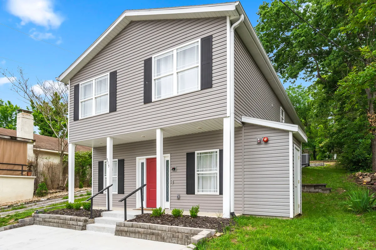 An inviting exterior view highlighting the red front door and symmetrical windows, framed by lush greenery. The porch provides a gathering spot, and the overall scene conveys relaxation and togetherness amid a vibrant neighborhood atmosphere.