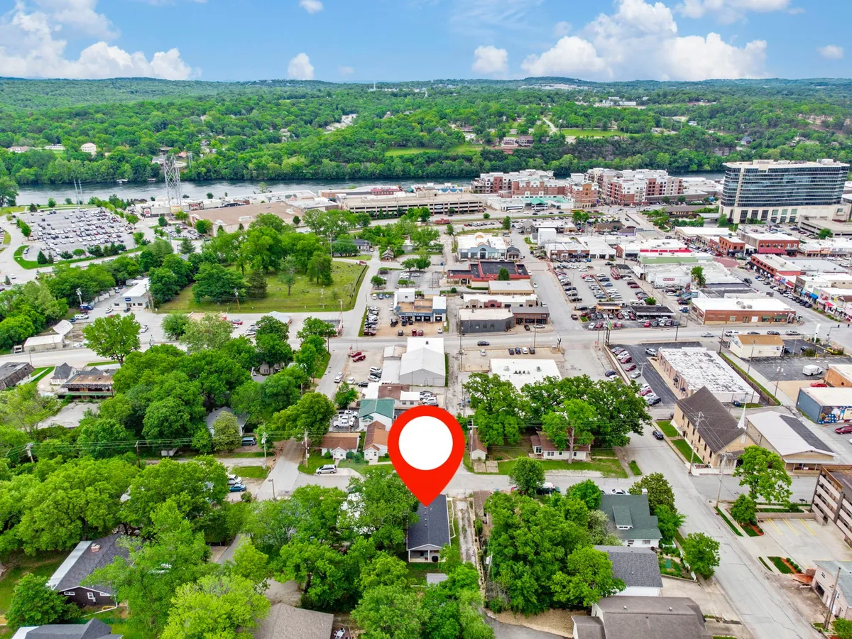 A vibrant exterior shot showcasing the highlighted property nestled near a lively riverfront, surrounded by greenery and nearby commercial buildings. The scene captures outdoor activity, with visible walking paths, the flowing river, and a glimpse of local shops and event spaces.