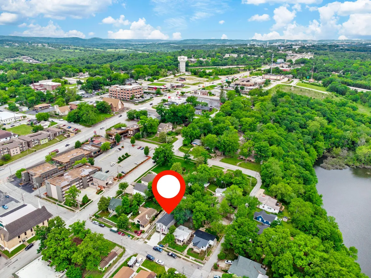 An inviting neighborhood scene featuring residential homes shaded by green trees, a main road, and a river alongside the community. People can be imagined walking, biking, or picnicking by the water, evoking both exploration and relaxation.