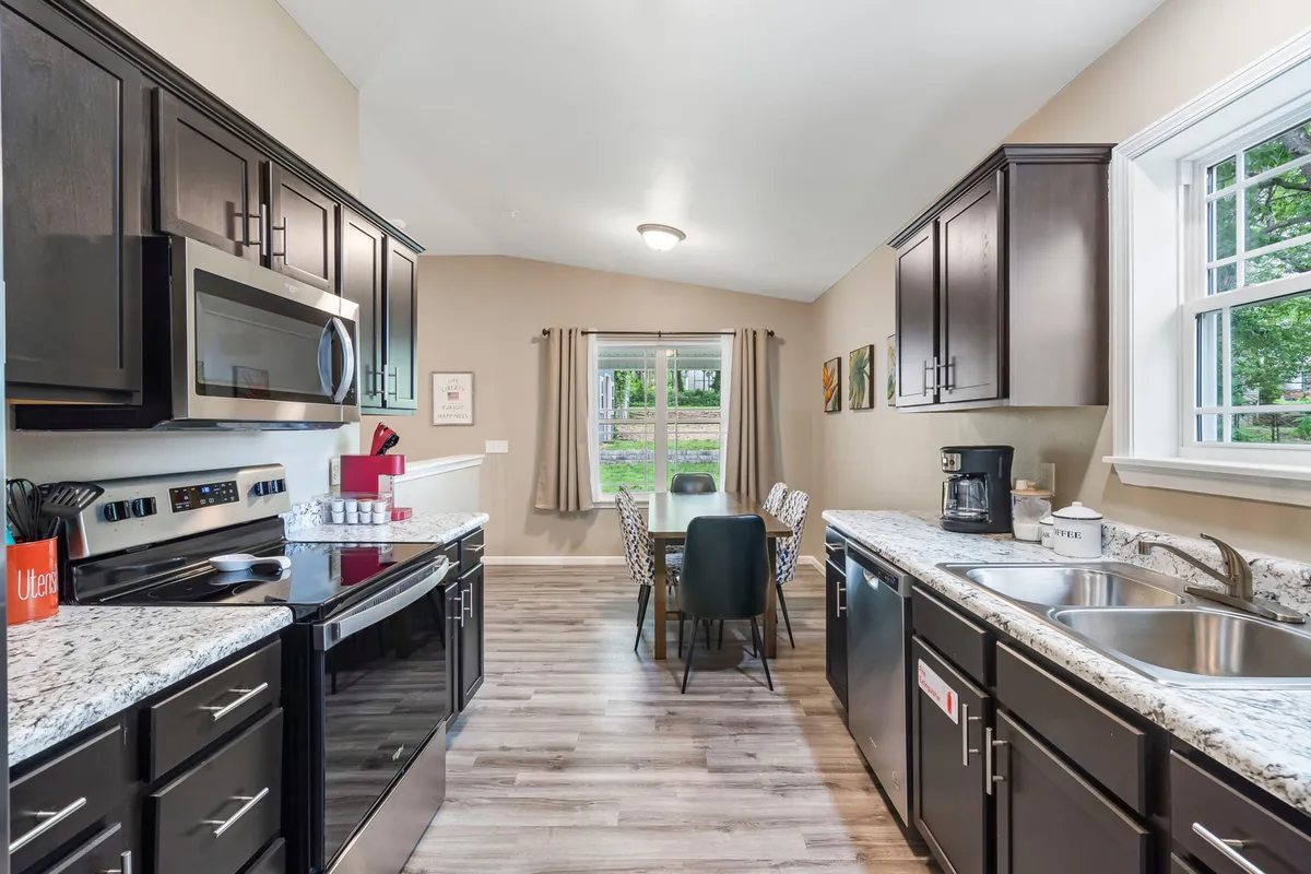 This image captures a welcoming, open-plan area featuring a granite countertop kitchen with black cabinetry, seamlessly flowing into a dining table with chairs set near a window. The scene is bathed in warm, natural light and exudes a sense of communal energy, with spaces clearly designed for cooking, dining, and socializing.
