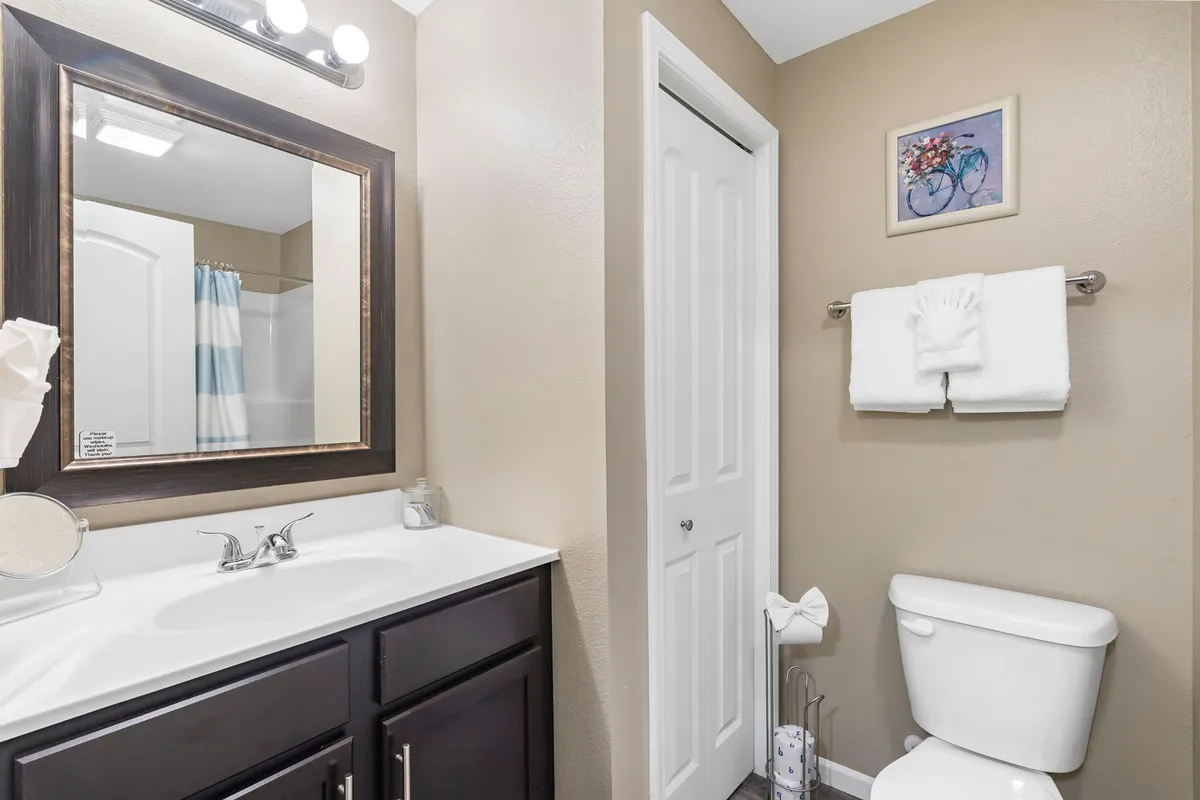 A fresh, inviting bathroom featuring a well-lit vanity with a sink, mirror, toilet, towel rack, and decorative artwork. The space appears spotless and thoughtfully designed for guest comfort.