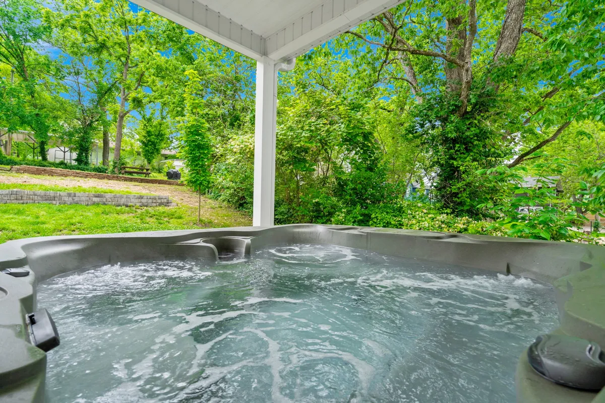 The image showcases the exterior of a charming single-family house surrounded by lush greenery. A spacious patio with comfortable seating is the focal point, featuring a private hot tub perfectly positioned for enjoying sunset views. The setting feels serene and inviting, with hints of nature all around, ideal for relaxation and gathering with friends or family.