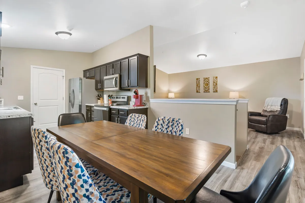 This inviting interior features a dining table set for a group, with kitchen appliances visible in the background and cozy, homely touches like wall art and a reclining chair. The space feels comfortable and designed for lingering over meals or friendly gatherings.